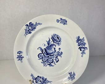 May include: A white ceramic plate featuring a central blue floral pattern and smaller blue floral motifs arranged around the rim. The plate is displayed on a gold-toned metal stand, showcasing a classic circular shape with a smooth, glazed finish.