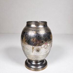 Puede incluir: Un jarrón de metal plateado con cuerpo redondeado y base de pedestal. La superficie presenta líneas horizontales grabadas y motivos florales con pequeños detalles circulares rojos. El metal muestra signos de envejecimiento y desgaste.