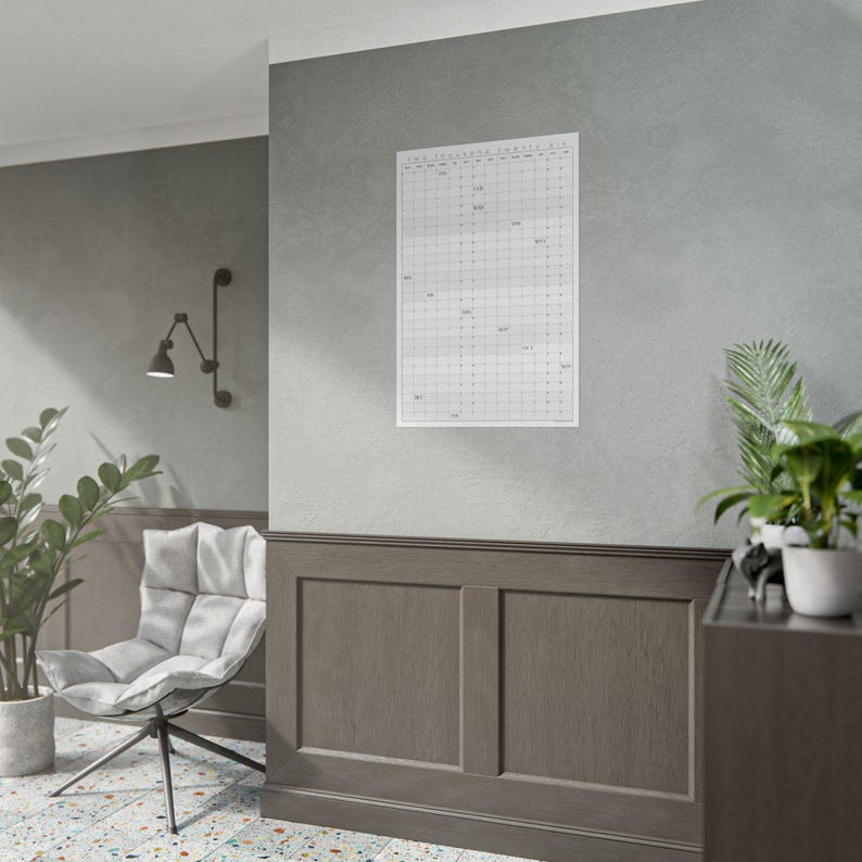 Op de afbeelding: Een witte rasterkalender met tekst is aan een grijze muur bevestigd. De kalender is rechthoekig en heeft een eenvoudig ontwerp. De kamer heeft een moderne uitstraling, met een grijze stoel, een zwarte lamp en potplanten.