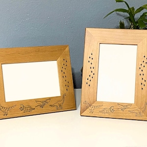 May include: Two wooden picture frames with a light brown finish. Each frame has a white interior and is decorated with dinosaur silhouettes and footprints. The frames are rectangular and suitable for displaying photos or artwork.