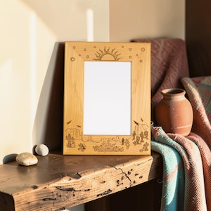 May include: A rectangular light-colored wooden picture frame featuring laser-engraved desert-themed illustrations. The top border displays a sun and stars, while the bottom border depicts a landscape with various cacti and rock formations.