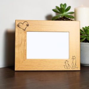 May include: A rectangular light brown wooden picture frame with a horizontal orientation. The top left corner features a laser-engraved heart shape formed by two paw prints. The bottom right corner displays a line art illustration of two cats.