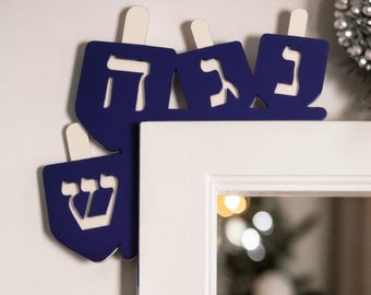 May include: A dark blue wooden door corner decoration featuring four dreidel shapes. Each dreidel is cut out with a different Hebrew letter in white, including Nun, Gimel, Hey, and Shin. The piece is designed to fit snugly around the corner of a door frame.