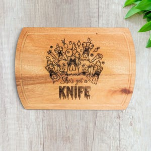May include: A rectangular wooden cutting board with rounded corners and a grooved border. The center features a laser-engraved illustration of various farm animals with wide-eyed expressions and the text "She's got a KNIFE" in a bold font.
