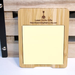 May include: A rectangular bamboo notepad holder with a light yellow sticky note pad. The holder has a carved design at the top with the text "Laser Made Gifts" and a website address. The notepad holder is next to a wooden crate.