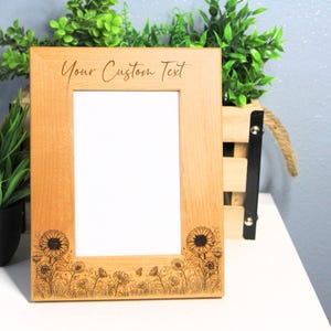 May include: A light-colored wooden picture frame with a rectangular opening. The frame is decorated with a sunflower and wildflower design. The text "Your Custom Text" is engraved at the top. The frame is on a white surface.