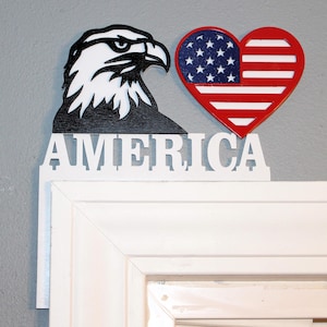 May include: A patriotic wall hanging featuring a black and white eagle head, a red, white, and blue heart-shaped American flag, and the word "AMERICA" in white letters. The design is mounted on a white door frame.