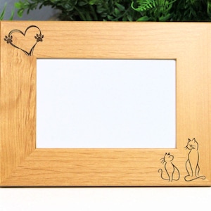 May include: A light brown wooden picture frame with a rectangular opening. The frame features etched designs of a heart with paw prints and two cats. The frame is displayed in front of greenery.