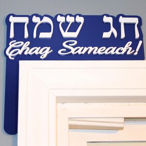 May include: A blue sign with white lettering that says "Chag Sameach!" in English and Hebrew. The sign is mounted above a white door frame.