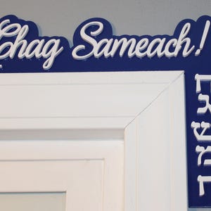 May include: A blue and white wooden sign with the words "Chag Sameach!" in white lettering. The sign also has Hebrew lettering in white on a blue background.