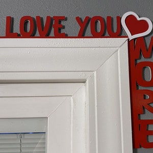 May include: Red wooden letters spelling "I LOVE YOU MORE" with a white heart in the middle of the "YOU" letters. The letters are attached to a white door frame.