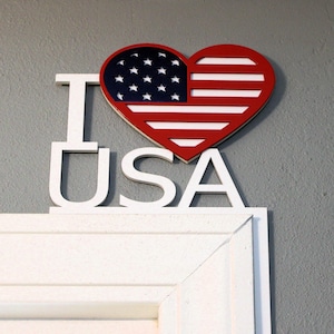 May include: A wooden sign with the words "I LOVE USA" in white. The heart-shaped "LOVE" section features the American flag design in red, white, and blue. The sign is mounted on a gray wall above a white door frame.