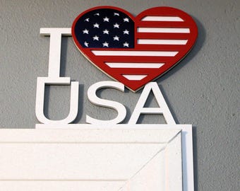 以下が含まれることがあります： 「I LOVE USA」の白い木製サイン。 「LOVE」のハートはアメリカ国旗のデザインで、赤と白のストライプと白い星の青い部分があります。 このサインは、白いドアフレームの上にある灰色の壁に取り付けられています。