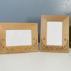 May include: Two wooden picture frames with floral corner designs. The frames are rectangular with a light brown finish and white interiors. One frame is horizontal, the other vertical. The frames are displayed on a white surface.