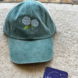 May include: Teal baseball cap with embroidered light blue and green floral design. A small blue square with white text that reads "ORB DESIGNS EST. 2023" is next to the cap.