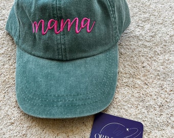 May include: A green baseball cap with the word "mama" embroidered in pink script. The cap has a curved brim and a fabric strap. A small, square, purple tag with white text that reads "ORB DESIGNS EST. 2023" is also present.