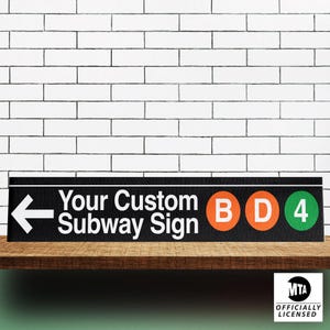May include: A black rectangular sign with white text that reads "Your Custom Subway Sign". The sign features a white arrow, and orange and green circles with the letters "B", "D", and the number "4". The sign is officially licensed by the MTA.
