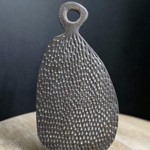 Peut inclure: Une râpe en céramique gris anthracite foncé en forme de goutte d'eau avec une poignée circulaire sur le dessus. La surface présente un motif dense et uniforme de petites indentations verticales en relief, avec une finition mate.