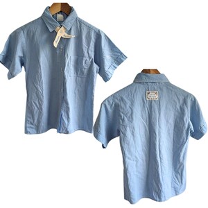 May include: Pale blue short-sleeved shirt with a button-down front, a chest pocket, and a collar. The shirt has a tag attached to the collar. The back of the shirt is also visible. The shirt is hanging on a wooden hanger.
