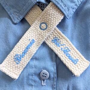 May include: Close-up of a light blue button-down shirt with two cream-coloured knitted collar stays. The collar stays have blue embroidered text that reads "Brownie" and "Girl Scouts". A blue embroidered circle is on one stay.