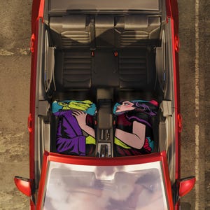 May include: Overhead view of a red convertible car interior with two black leather seats. Two headrest pillows feature comic book style illustrations. One shows a green-skinned character in a purple robe, the other a woman with dark hair.