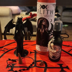 May include: A red fabric cloth featuring a black sigil, a black winged figurine, a tall red candle with a Lilith label, a black spray bottle with a moon charm, a small brown incense cone, a black cord bracelet, and a small brown stone.