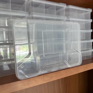 Puede incluir: Contenedores de almacenamiento de plástico transparente con múltiples compartimentos y tapas. Los contenedores están apilados en un estante de madera. Las tapas tienen un diseño rectangular con una pequeña zona perforada.