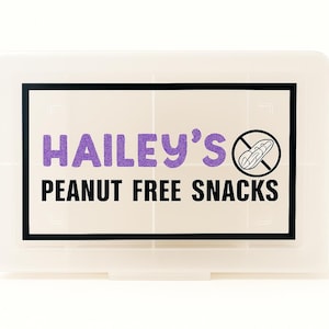 May include: A white plastic container with a black border and the text "Hailey's Peanut Free Snacks" in purple and black. A peanut with a slash through it is in the upper right corner.