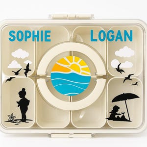 May include: A beige lunchbox with multiple compartments, featuring the names "SOPHIE" and "LOGAN" in blue. The design includes a sun and waves graphic, clouds, seagulls, and beach-themed silhouettes. The lunchbox has a clear lid and a white clasp.