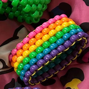 Könnte beinhalten: Ein regenbogenfarbenes Perlenarmband mit lila, blauen, grünen, gelben, orangen und rosa Perlen. Das Armband besteht aus kleinen, runden Perlen, die in einem sich wiederholenden Muster aufgereiht sind.