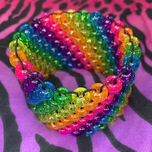 May include: A wide cuff bracelet constructed from multiple rows of translucent, multi-coloured plastic pony beads in shades of red, orange, yellow, green, blue, and purple, all strung together on thin black elastic cord in a repeating pattern.