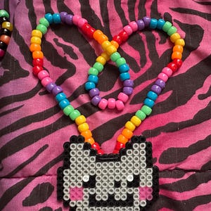 May include: A colorful beaded necklace with a rainbow-colored strand and a pendant shaped like a cat's face. The cat pendant is made of white, black, and pink beads. The necklace is displayed on a pink and black zebra-striped background.