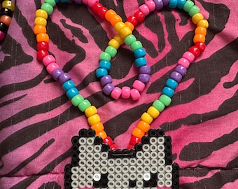 May include: A beaded necklace featuring a rainbow-coloured strand and a cat-shaped pendant. The pendant is made of white, black, and pink beads. The necklace is displayed against a pink and black zebra-striped background.