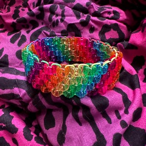 Könnte beinhalten: Ein lebendiges, regenbogenfarbenes Armband aus transparenten, quadratischen Perlen. Die Perlen sind in einem geschichteten Muster angeordnet und zeigen ein Farbspektrum. Das Armband liegt vor einem pinkfarbenen und schwarzen Tierdruck-Hintergrund.