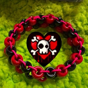 Puede incluir: Un brazalete de cuentas rojas y negras con un diseño de calavera y huesos cruzados dentro de un corazón rojo. El brazalete está hecho de pequeñas cuentas redondas.