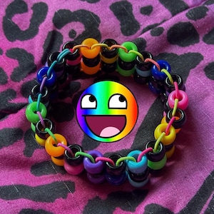 Könnte beinhalten: Ein farbenfrohes Kandi-Armband mit einem Regenbogen-Smiley in der Mitte. Das Armband besteht aus schwarzen, orangefarbenen, gelben, grünen, blauen, lila und rosa Perlen. Der Hintergrund ist ein pinkfarbener Stoff mit schwarzem Leopardenmuster.