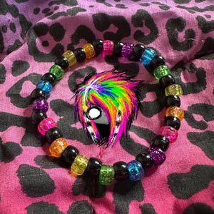 Può includere: Un braccialetto di perline arcobaleno con perline nere. Il braccialetto è su un tessuto a stampa leopardata rosa e nera.