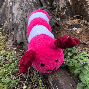 Könnte beinhalten: Ein rosa-weißes Häkeltier in Form einer Raupe mit schwarzen Augen und einem lächelnden Mund. Das Spielzeug liegt auf einem Baumstamm mit grünen Pflanzen im Hintergrund.
