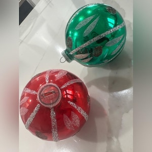 Pode incluir: Duas bolas de Natal vintage. Uma é vermelha com detalhes em glitter prateado e a outra é verde com detalhes em glitter prateado. Ambas são redondas e têm um acabamento brilhante, perfeitas para a decoração festiva.