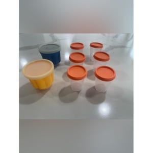 Pode incluir: Uma coleção de recipientes de plástico com tampas. Os recipientes são transparentes com tampas laranja. Há um recipiente azul com uma tampa cinza e um recipiente amarelo com uma tampa bege. Os recipientes estão em uma superfície branca.