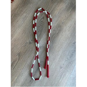 May include: A jump rope featuring a long cord with alternating red and white cylindrical segments. The rope includes two solid red cylindrical handles at one end, designed for gripping during use, with a consistent color pattern throughout the cord.