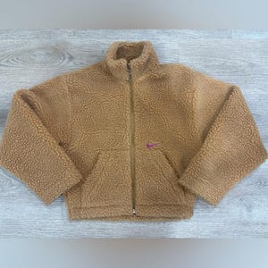 May include: A tan, long-sleeved, full-zip jacket made of a textured, fuzzy sherpa fleece material. The jacket features a high collar, a central zipper closure, and a small, embroidered purple Nike swoosh logo on the left side of the chest.