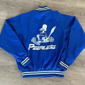 May include: Royal blue jacket with white and navy stripes on the collar, cuffs, and hem. The back features a cartoon duck graphic with a baseball bat and the word "Peerless" in white block letters. A vintage-style athletic jacket.