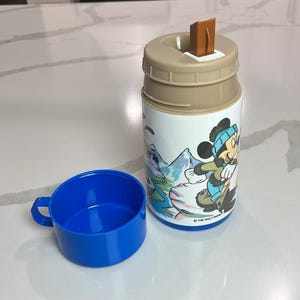 May include: A vintage Disney thermos and cup set. The thermos features a cartoon Mickey Mouse design in winter clothing, with a tan lid and brown flip-top. The cup is bright blue. The thermos has a white body with a cartoon image of Mickey Mouse.