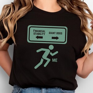 May include: Black t-shirt with a green graphic of a person running away from a sign that says "Financial Stability" with an arrow pointing left and "Giant Dogs" with an arrow pointing right. The text "ME" is below the running person.
