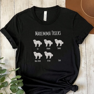 Pode incluir: Camiseta preta com um gr&aacute;fico branco de um c&atilde;o da ra&ccedil;a Maremma. O gr&aacute;fico mostra o c&atilde;o realizando diferentes truques, incluindo sentar, deitar, sacudir, rolar, buscar e ficar quieto. O texto "Maremma Tricks" est&aacute; acima do gr&aacute;fico.
