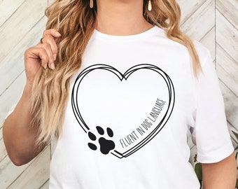Może przedstawiać: Biała koszulka z czarnym wzorem w kształcie serca, odciskiem łapy i napisem "FLUENT IN DOG LANGUAGE". Koszulka wykonana jest z miękkiego materiału i ma swobodny krój.