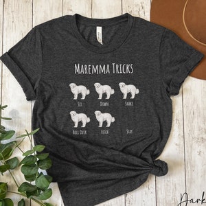 Pode incluir: Camiseta cinza escuro heather com uma ilustra&ccedil;&atilde;o de um c&atilde;o Maremma branco e o texto "Maremma Tricks". O c&atilde;o &eacute; mostrado em diferentes poses, com o truque correspondente abaixo de cada pose: sentar, deitar, sacudir, rolar, buscar, ficar.