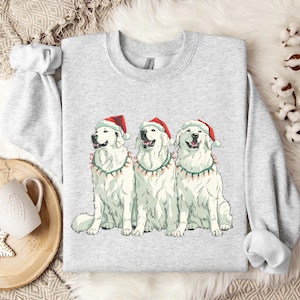 Puede incluir: Una sudadera gris claro con un diseño de tres perros blancos con gorros de Papá Noel y luces navideñas. Los perros sonríen y están sentados uno al lado del otro. La sudadera tiene cuello redondo y mangas largas.