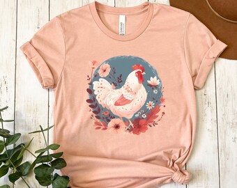 May include: A peach-colored t-shirt with a graphic of a white chicken surrounded by pink and red flowers on a blue background. The text "Heather Peach" is printed on the bottom right corner of the shirt.